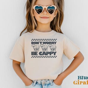 May include: A young person wearing a light beige t-shirt with a graphic of three capybaras and the text "Don't Worry Be Cappy". The shirt has a black and white checkered pattern around the graphic. The person is wearing heart-shaped sunglasses.