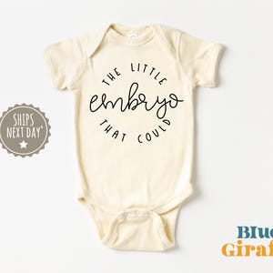 Può includere: Un body per neonato color crema con testo nero che recita "The little embryo that could".