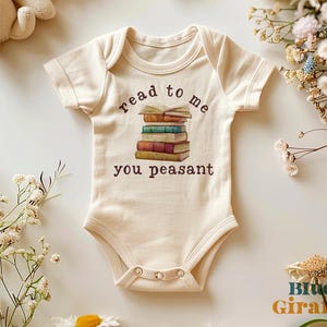May include: Cream-coloured baby onesie with short sleeves. The onesie features a graphic of a stack of books with the words "read to me you peasant" printed in brown. The onesie has three snaps at the bottom.