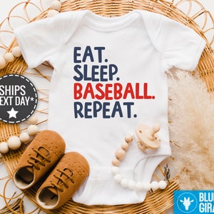 May include: White baby onesie with blue text that reads "Eat. Sleep. Baseball. Repeat." The word "Baseball" is in red.