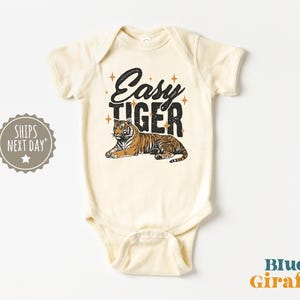 K&ouml;nnte beinhalten: Cremefarbener Baby-Body mit dem Text "Easy Tiger" in schwarzer Schrift &uuml;ber einer Tiger-Illustration. Der Tiger ist orange und schwarz. Der Body hat kurze &Auml;rmel und einen Druckknopfverschluss.