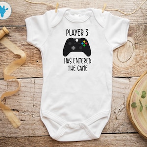 Puede incluir: Un body blanco para bebé con texto negro que dice "Player 3 has entered the game". Un gráfico negro de un mando de videojuego está por encima del texto.