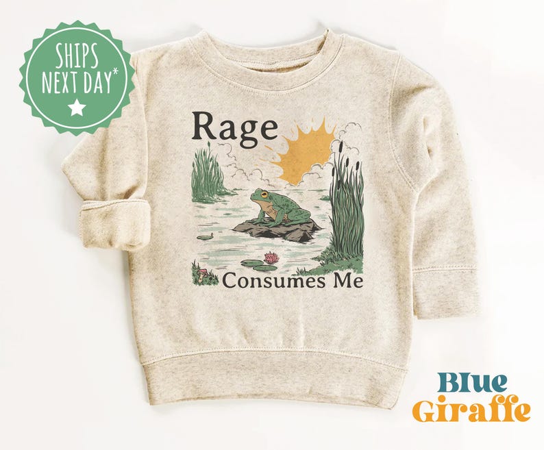 Puede incluir: Una sudadera beige de cuello redondo con un estampado gr&aacute;fico de una rana verde sentada sobre una roca en un estanque con juncos y un sol amarillo. El texto "Rage" est&aacute; impreso sobre la rana y "Consumes Me" debajo de la escena.