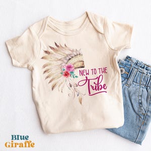 Puede incluir: Un body de bebé de manga corta en color crema con un gráfico de un tocado de plumas adornado con flores rosas y rojas. El texto "NEW TO THE TRIBE" está impreso en una fuente de color rosa oscuro, tipo script y sans-serif, junto al dibujo.