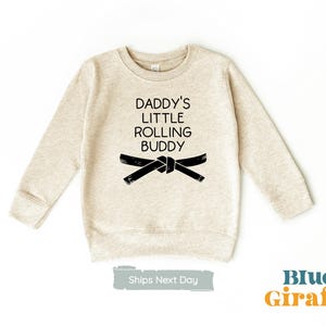 May include: A beige sweatshirt with black text that reads "Daddy's Little Rolling Buddy" and a black belt graphic.