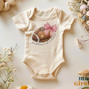 May include: A cream-coloured baby onesie with a brown football and a pink bow. The text "huddles and cuddles" is printed on the onesie.
