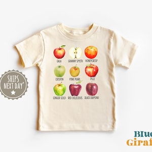 May include: Cream-colored t-shirt featuring watercolor illustrations of various apple varieties, including Gala, Granny Smith, and Honeycrisp. The shirt also displays the text "Blue Giraffe" and "Ships Next Day".