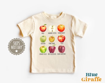May include: Cream-colored t-shirt featuring watercolor illustrations of various apple varieties, including Gala, Granny Smith, and Honeycrisp. The shirt also displays the text "Blue Giraffe" and "Ships Next Day".