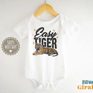 K&ouml;nnte beinhalten: Wei&szlig;er Baby-Body mit dem Text "Easy Tiger" in schwarzer und goldener Schrift, mit einer Tiger-Illustration. Der Body h&auml;ngt an einem Holzb&uuml;gel.