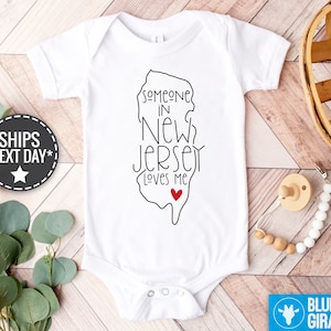May include: White baby onesie with a black outline of the state of New Jersey and the text "Someone in New Jersey loves me" with a red heart.