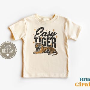 K&ouml;nnte beinhalten: Cremefarbenes Kinder-T-Shirt mit einem Tiger-Motiv und dem Schriftzug "Easy Tiger" in Schwarz. Der Tiger ist mit braunen und schwarzen Streifen dargestellt. Das T-Shirt hat kurze &Auml;rmel.