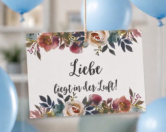 May include: A rectangular white sign hanging from a string, featuring a watercolor floral border in shades of burgundy, cream, and dark green. The text "Liebe liegt in der Luft!" is printed in a black script font in the center of the sign.