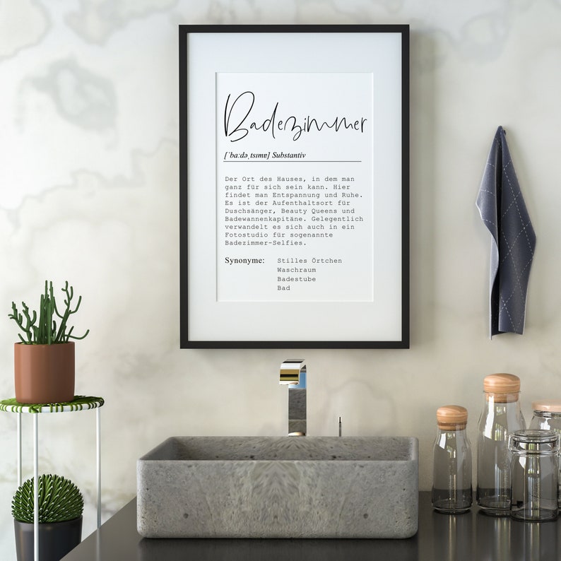 May include: A black framed print with the word "Badezimmer" in white text. The print is hanging on a white wall above a gray rectangular sink with a chrome faucet. There are two small glass jars with wooden lids on the counter to the right of the sink.