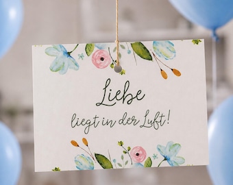 May include: A rectangular white card hanging from a thin string, featuring a watercolor floral border with blue and pink flowers and green leaves. The text "Liebe liegt in der Luft!" is printed in dark green cursive and sans-serif lettering.