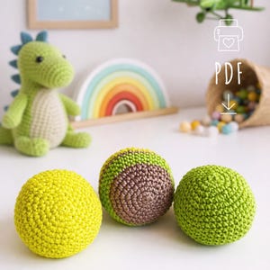 Puede incluir: Tres bolas de ganchillo en amarillo, verde y marrón, con un dinosaurio verde y un arcoíris en el fondo. Las bolas están hechas a mano y parecen ser para niños. La imagen tiene una estética suave y acogedora.