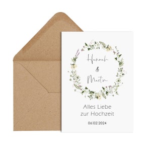 May include: A white card with a floral wreath design. The wreath is made of green leaves and yellow flowers. The text "Hannah & Martin" is in the center of the wreath. The text "Alles Liebe zur Hochzeit" and the date "06/02/2024" are below the wreath.