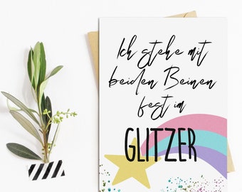 May include: A white card with a rainbow and a gold star. The text on the card reads "Ich stehe mit beiden Beinen fest im GLITZER".