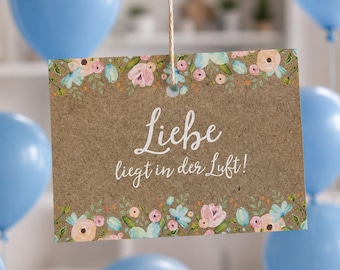May include: A rectangular brown kraft paper sign hanging from a string, featuring a floral border of pink, peach, and light blue flowers. The text "Liebe liegt in der Luft!" is printed in white script in the center, surrounded by light blue balloons.