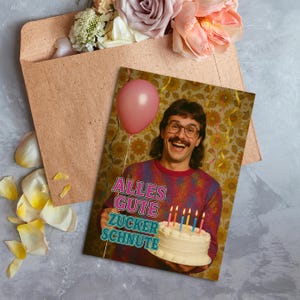 May include: A birthday greeting card featuring a man holding a cake with lit candles and a pink balloon. The card has the text "ALLES GUTE ZUCKER SCHNUTE". The card is next to a brown envelope and flowers.
