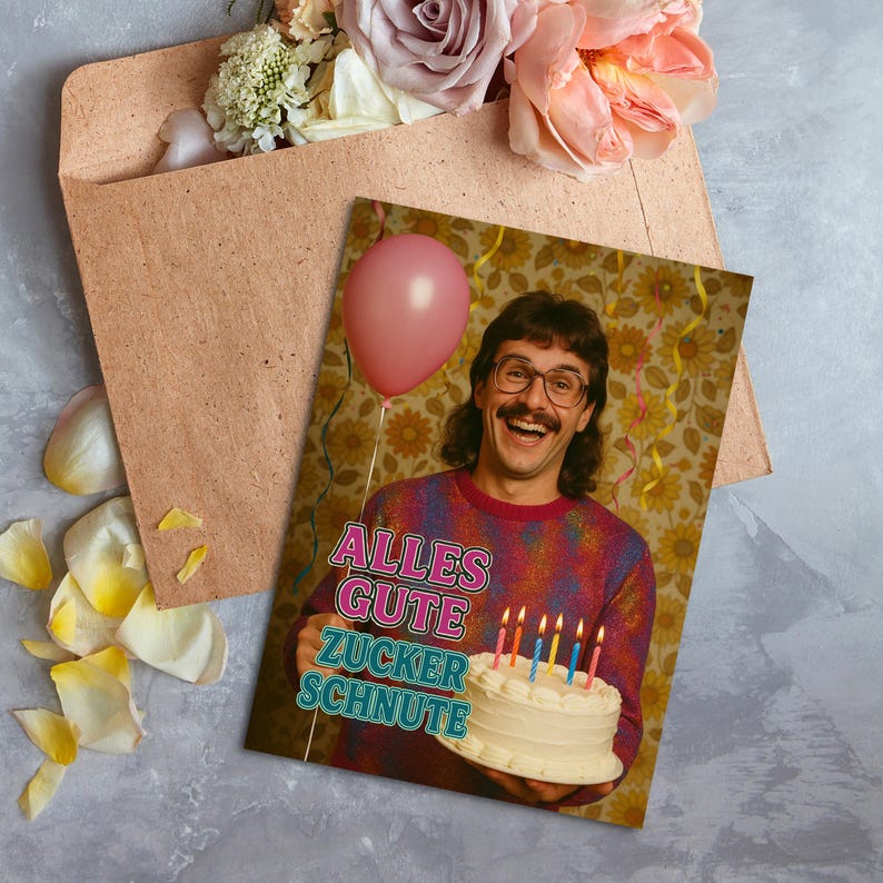 May include: A birthday greeting card featuring a man holding a cake with lit candles and a pink balloon. The card has the text "ALLES GUTE ZUCKER SCHNUTE". The card is next to a brown envelope and flowers.