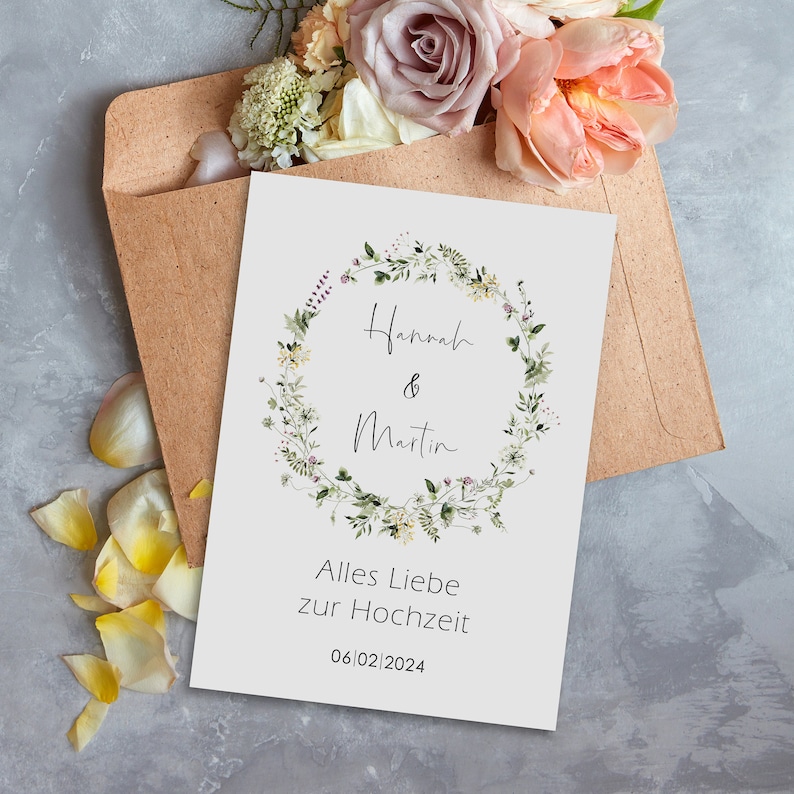 May include: A white card with a floral wreath design and the text "Hannah & Martin" in black script. Below the wreath is the text "Alles Liebe zur Hochzeit" and the date "06/02/2024".