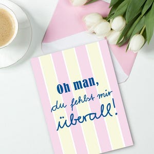 Puede incluir: Una tarjeta de felicitación con rayas verticales rosas y amarillas, con el texto "Oh man, du fehlst mir überall!" en escritura azul. La tarjeta está acompañada de un sobre rosa, una taza de café y tulipanes blancos.