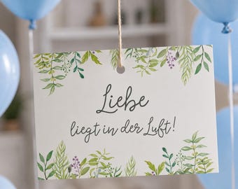 May include: A rectangular white card hanging from a string, featuring a watercolor botanical border of green leaves and small purple berries. The text "Liebe liegt in der Luft!" is printed in a dark gray script font in the center of the card.