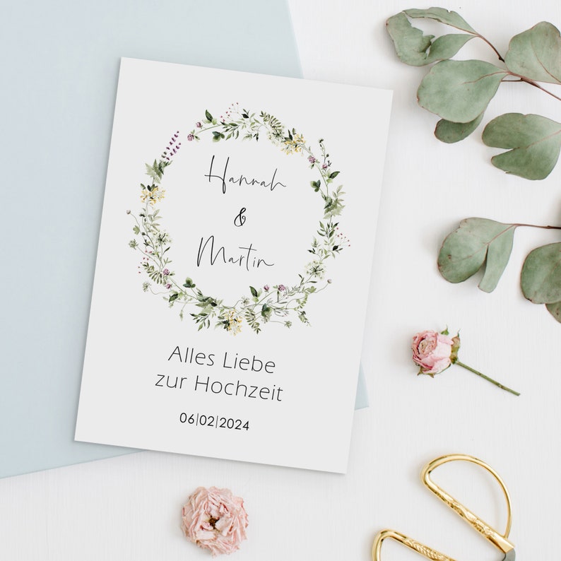 May include: A white card with a floral wreath design. The wreath is made of green leaves and small yellow and pink flowers. The text "Hannah & Martin" is written in black cursive inside the wreath. Below the wreath, the text "Alles Liebe zur Hochzeit" and the date "06|02|2024" are written in black.