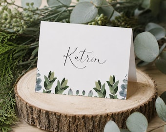 May include: A white folded place card featuring the name Katrin in black cursive script, decorated with a printed border of green eucalyptus leaves along the bottom edge, displayed on a circular wooden slice with natural bark edges.