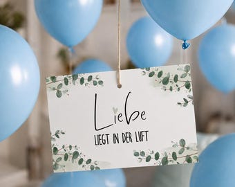 May include: A rectangular white sign hanging from a string, decorated with green eucalyptus leaf illustrations in the corners. The text reads "Liebe" with a small heart above the "i" and "LIEGT IN DER LUFT" below, surrounded by light blue balloons.