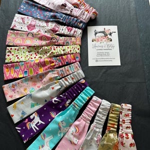 May include: A selection of patterned fabric headbands, featuring designs such as rainbows, ballerinas, and unicorns. The headbands have elasticated sections. A card with the text "Heavens 2 Bitsy" is also visible.