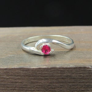 May include: A silver ring with a pink gemstone set in the center. The ring has a curved band.