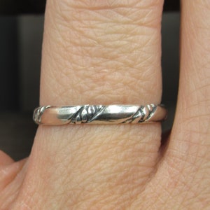 May include: A silver band ring with a textured design. The ring is worn on a finger.