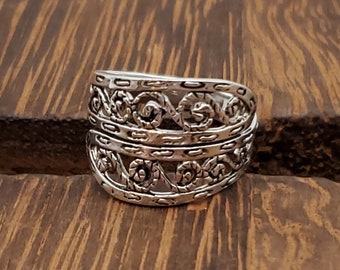 May include: A silver ring with a wide band featuring an intricate, ornate design of interwoven swirls and lines.