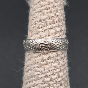 May include: A silver ring with an intricate pattern of repeating shapes and designs. The ring is displayed on a beige fabric finger stand against a dark gray background. The ring's design includes a central figure with symmetrical patterns.