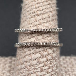 May include: Two silver rings with small, clear stones set in a row. The rings are displayed on a beige ring holder against a dark gray background. The rings are delicate and appear to be made of a shiny metal.