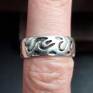 May include: A silver ring with a textured design resembling flames. The ring is worn on a finger.