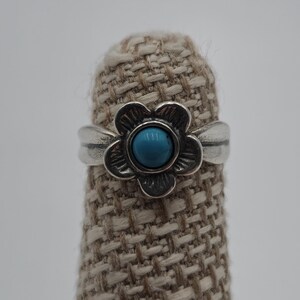 May include: A silver ring featuring a flower design with a turquoise center stone. The ring is set on a textured, tan surface against a white background. The flower petals are detailed, and the band is smooth.