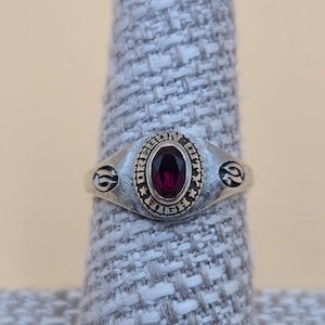 May include: A silver-toned ring with a dark red oval stone at the center. The ring has an oval face with the words "OREGON CITY" around the stone. The ring is on a gray fabric display.