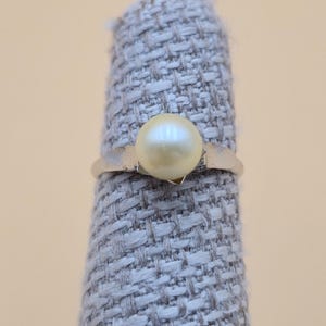 May include: A silver ring featuring a single, round, cream-colored pearl. The pearl is the focal point, set on a simple, thin band. The ring is displayed on a gray fabric surface against a neutral background.