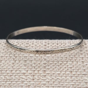 May include: A thin, circular silver-toned metal bangle bracelet featuring a delicate, continuous engraved pattern along its entire outer surface. The bracelet has a smooth, polished finish and a consistent, slender profile throughout its circumference.