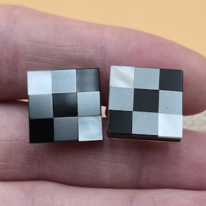 May include: A pair of square cufflinks with a checkerboard pattern. The pattern alternates between black, white, and gray squares. The cufflinks are set against a neutral background.