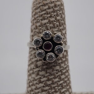 May include: A silver ring featuring a floral design. The ring has a central, round, dark red stone, surrounded by six clear, round stones. The ring is displayed on a beige, textured surface against a white background.