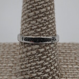 May include: A silver ring with the words "I am enough" engraved on it. The ring is displayed on a beige fabric ring holder.