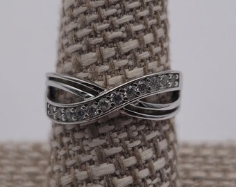 May include: A silver ring with an infinity symbol design. The ring has a row of small, clear stones set in the centre of the infinity symbol. The ring is displayed on a beige ring holder against a white background.