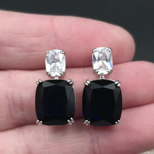 May include: A pair of square drop earrings with a silver setting. Each earring features a large, black, square-cut stone below a smaller, white, rectangular stone. The earrings are displayed against a neutral background.