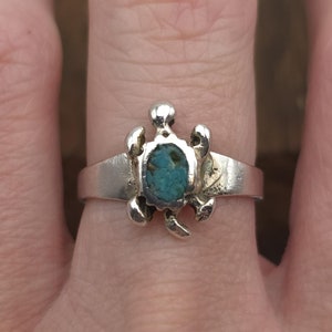 May include: A silver ring featuring a turtle design. The turtle has a turquoise stone set in its center. The ring is worn on a finger, showcasing the intricate details of the jewelry.