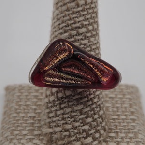May include: A triangular shaped ring with a red and gold glass inlay. The ring is on a fabric ring display stand.