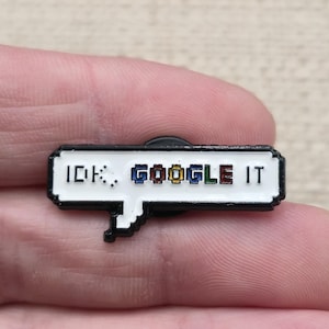 May include: A rectangular enamel pin shaped like a speech bubble with a white background and a black border. The text IDK, GOOGLE IT is printed in a pixelated font, with the word GOOGLE featuring blue, red, yellow, and green colored letters.