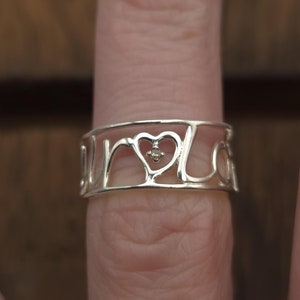May include: Silver ring with a heart design and the word "love" spelled out around the heart. The ring has a small diamond in the center of the heart.